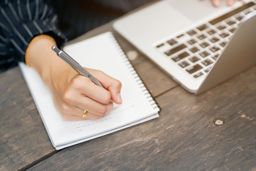 person writing on notebook next to a laptop