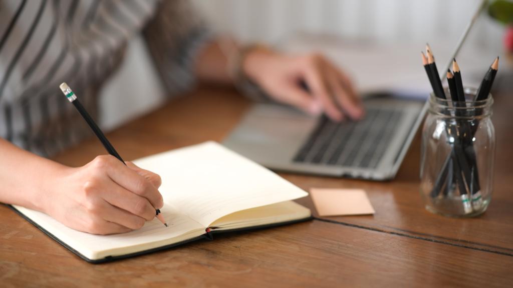 person writing on laptop and pad of paper