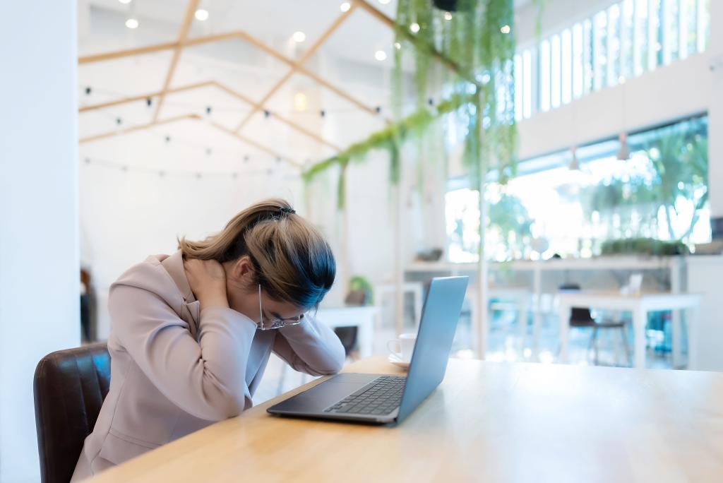 poor posture, painful neck concept - woman in a coffee shop with her laptop and looking down while pressing on her back and neck with her hands