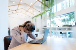 poor posture, painful neck concept - woman in a coffee shop with her laptop and looking down while pressing on her back and neck with her hands
