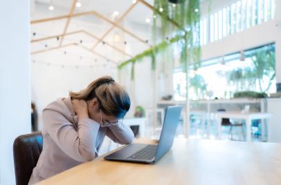 poor posture, painful neck concept - woman in a coffee shop with her laptop and looking down while pressing on her back and neck with her hands